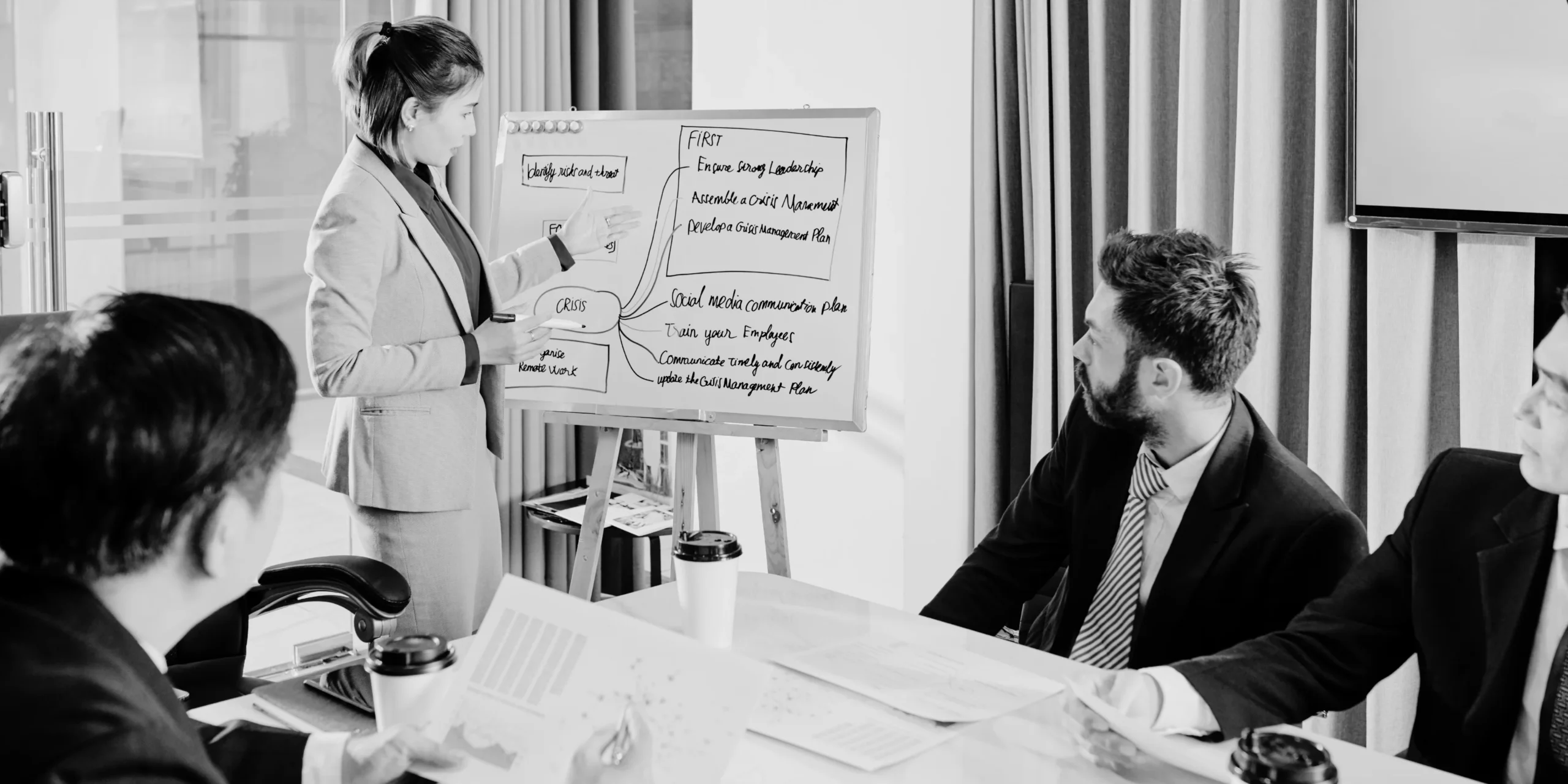 Ejecutiva explicando un esquema en una pizarra ante dos colegas sentados.