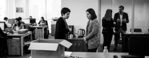 Dos trabajadoras conversan en voz baja junto a la zona del café mientras, al fondo, una jefatura habla en una sala de vidrio; en primer plano, una caja de cartón sobre un escritorio vacío.