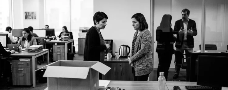 Dos trabajadoras conversan en voz baja junto a la zona del café mientras, al fondo, una jefatura habla en una sala de vidrio; en primer plano, una caja de cartón sobre un escritorio vacío.